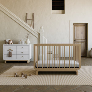 Light wood baby crib on patterned rug in open-concept room with matching two-tone white and light wood dresser behind it