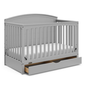 Convertible pebble gray crib with an open drawer, viewed from an angle
