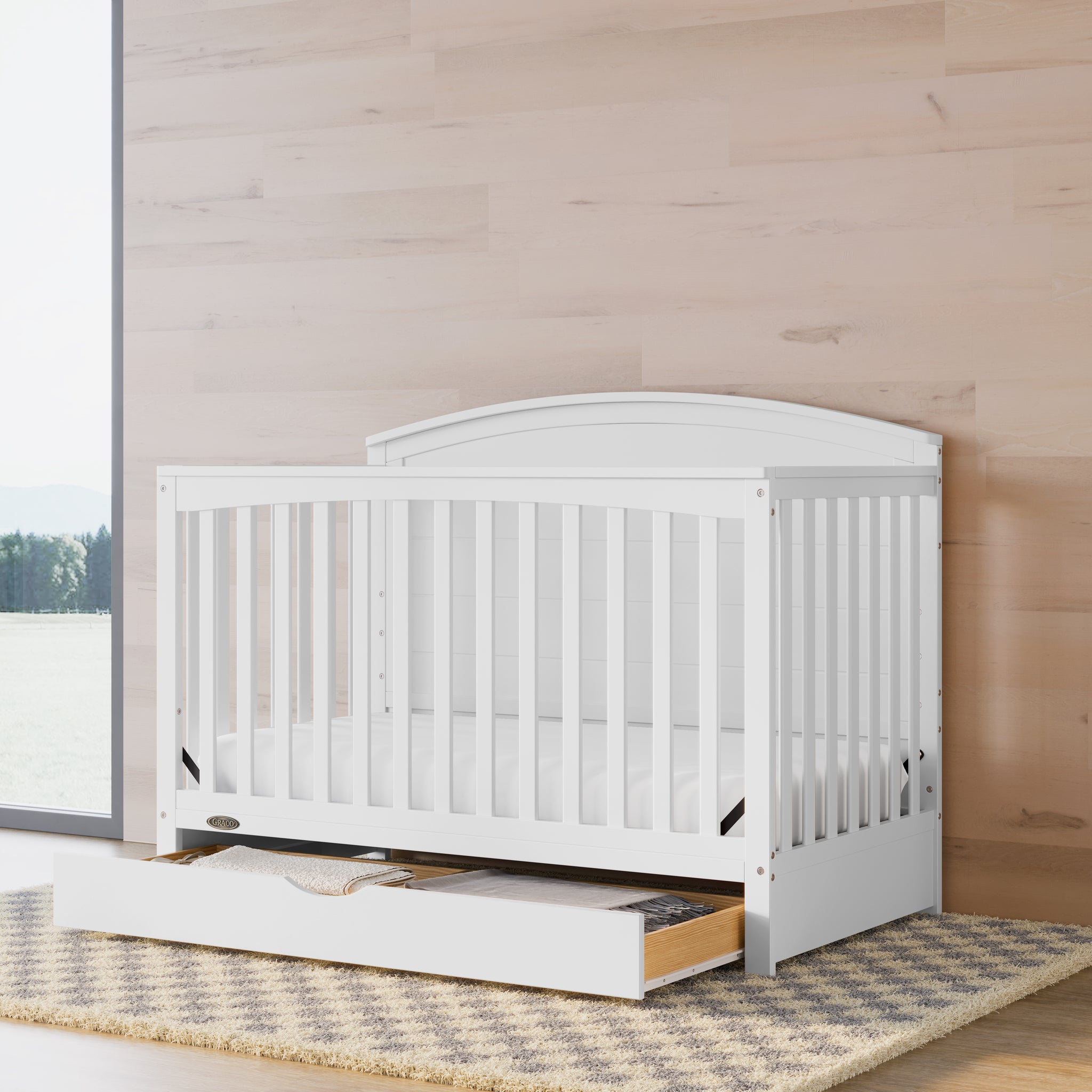 Convertible white crib with an open drawer in a nursery featuring wood-tone walls.