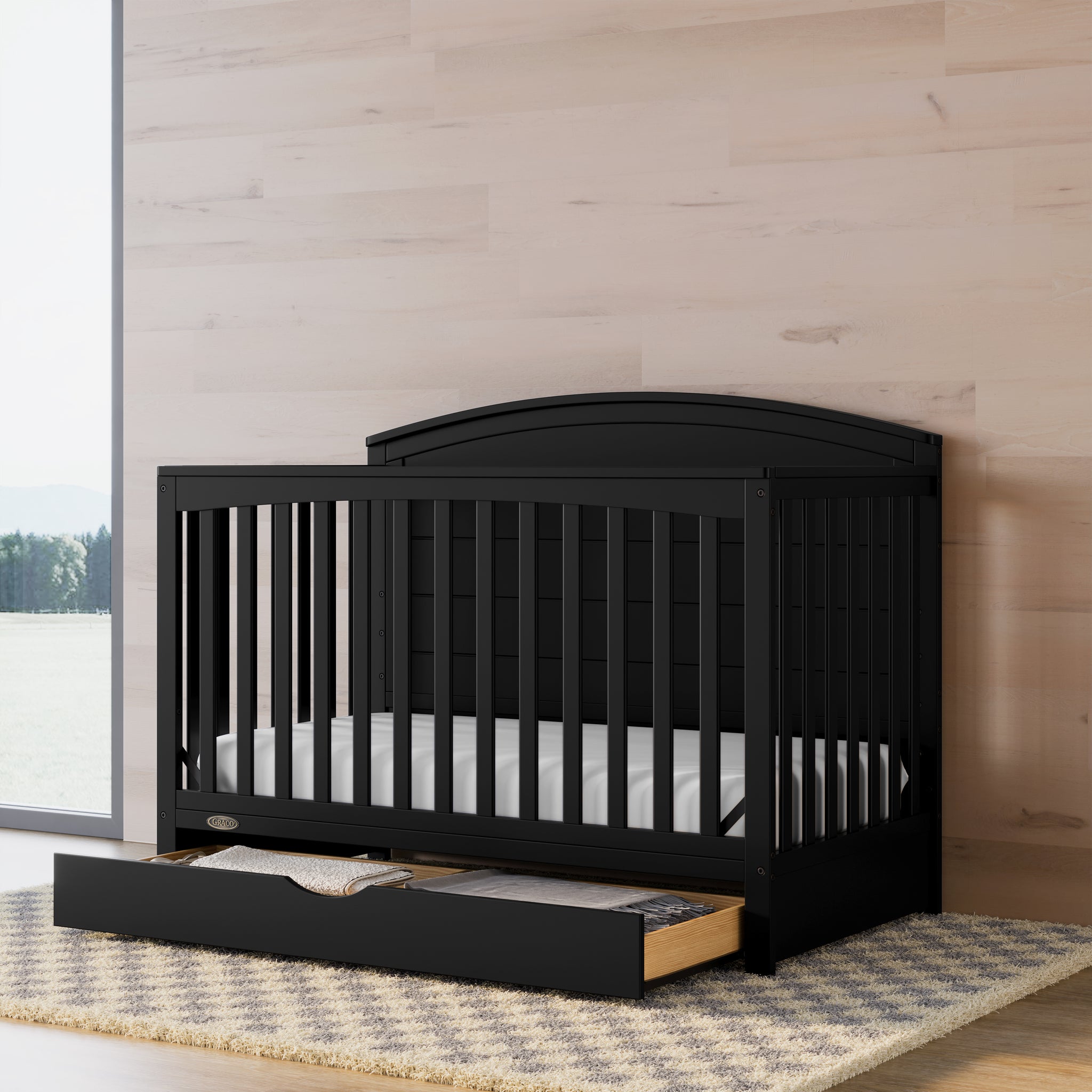 Convertible black crib with an open drawer in a nursery featuring wood-tone walls.