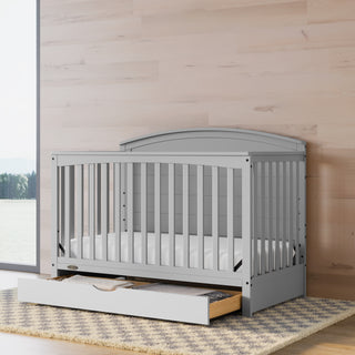 Convertible pebble gray crib with an open drawer in a nursery featuring wood-tone walls.