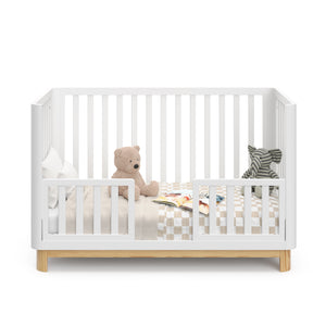 Front view of a white crib with a natural wood color base, featuring guard rails in toddler bed conversion