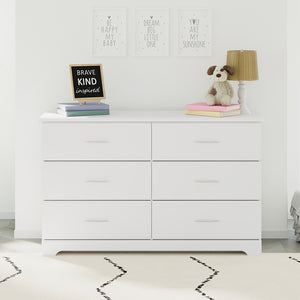 White 6 drawer double dresser in bedroom setting with picture frames above and various decorative objects and lamp on top of dresser