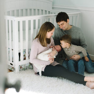 Mother holding baby, father and young son looking at baby, all sitting on floor of nursery in front of white crib