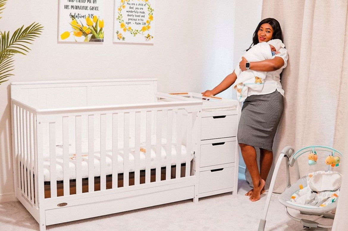 Woman holding baby in her arms, standing next to white crib in a nursery setting