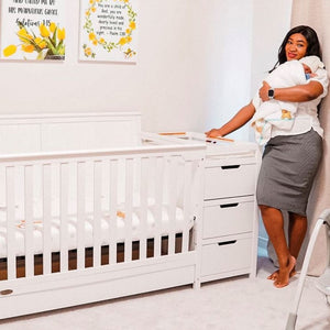 Woman holding baby in her arms, standing next to white crib in a nursery setting