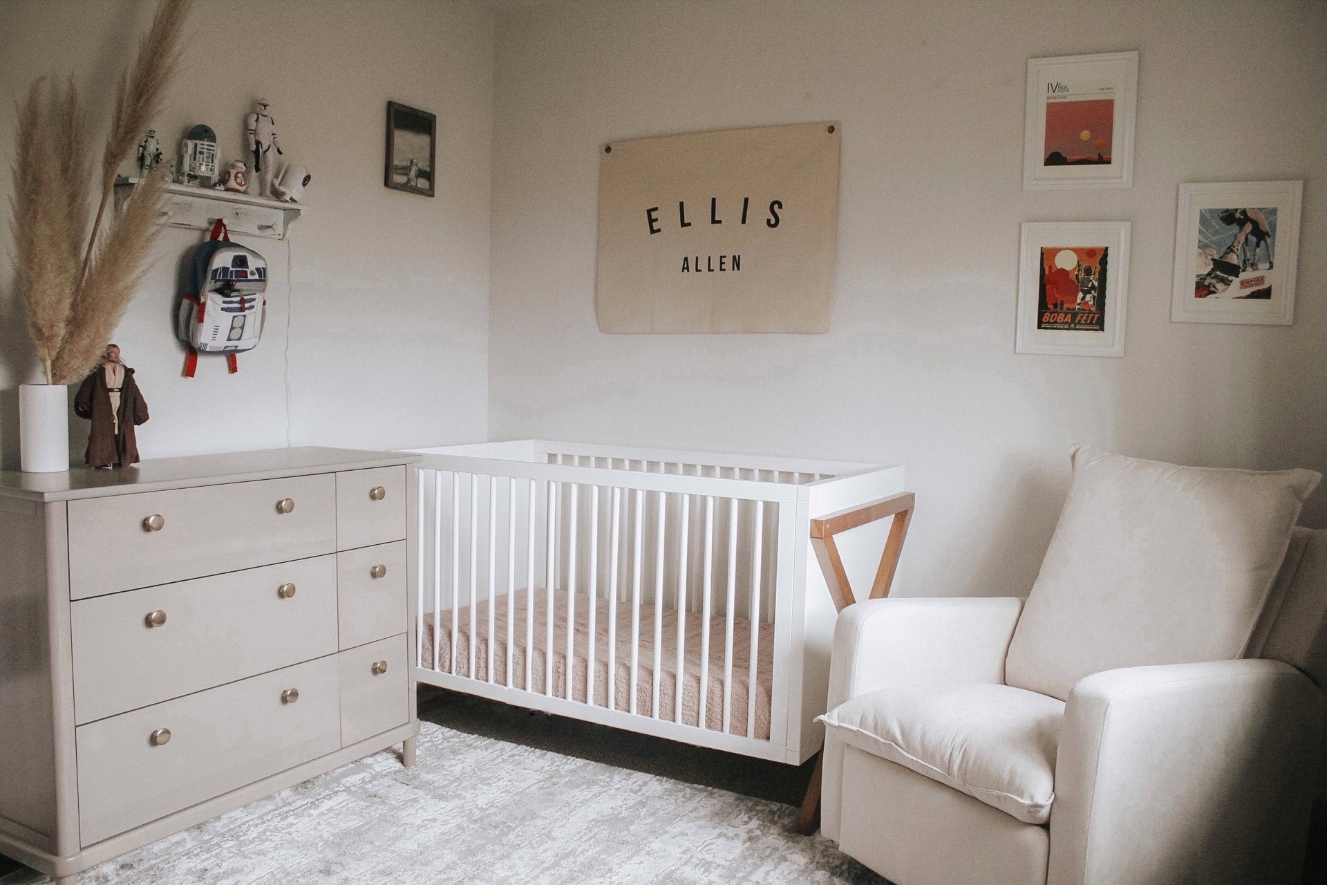 Nursery bedroom setting with beige dresser, white crib, and beige nursery chair. Room contains various decorative objects, with flag that reads Ellis Allen.