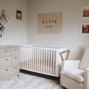 Nursery bedroom setting with beige dresser, white crib, and beige nursery chair. Room contains various decorative objects, with flag that reads Ellis Allen.