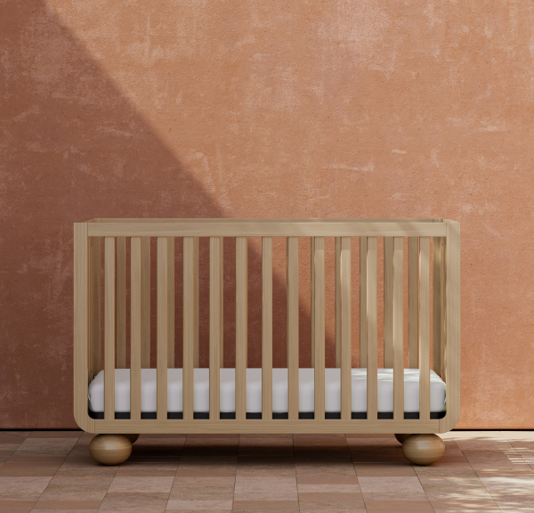 Natural light wood baby crib with round ball feet against terra cotta colored backdrop