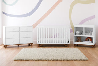 Room with shag rug prominently featured in the center, with white dresser, white crib, and white storage unit each with natural wood tone accent