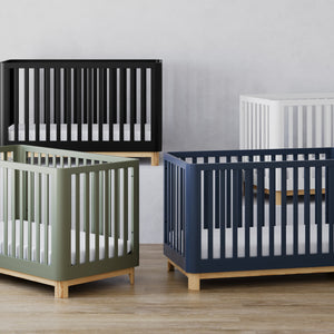 Studio setting with four baby cribs in various colors (black, white, olive, and navy blue) at various heights, each crib with a natural wood base