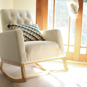 Beige upholstered nursery rocking chair with natural wood base, with multicolored cushion on chair, situated near window