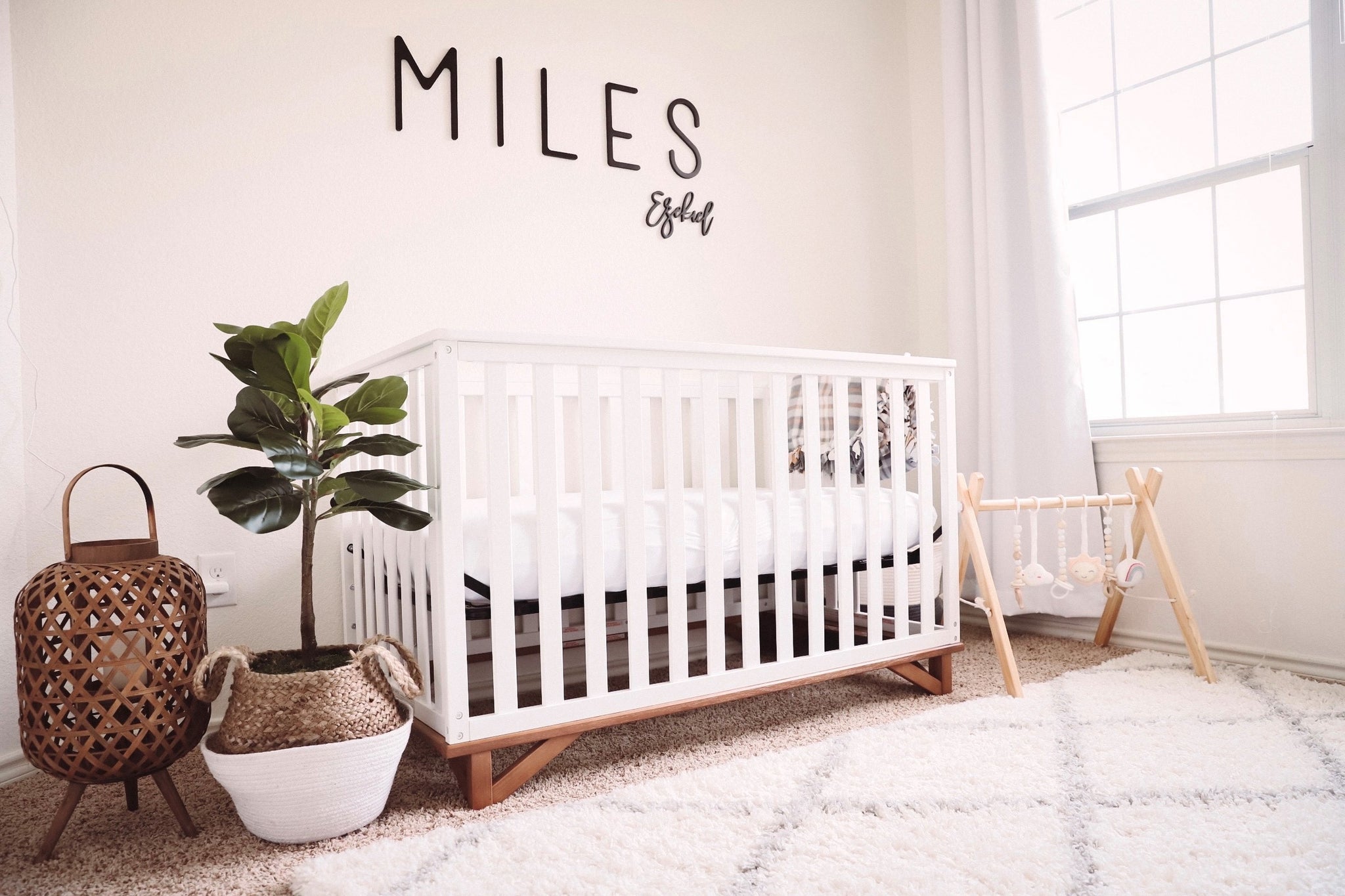 White crib with natural wood base situated against wall with words Miles Ezekiel displayed above crib, surrounded by various nursery decor