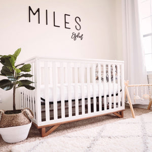 White crib with natural wood base situated against wall with words Miles Ezekiel displayed above crib, surrounded by various nursery decor