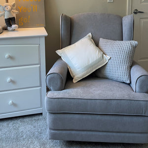 Gray chair in nursery bedroom setting, with two decorative pillows, with white dresser beside it, with various decorative objects around it.