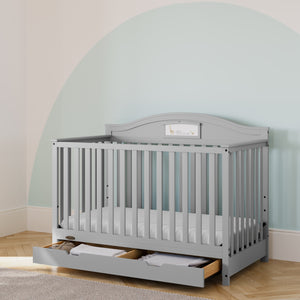 Light gray baby crib in room setting with under-crib drawer open