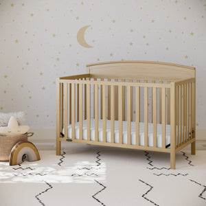 Natural wood colored baby crib in nursery bedroom setting, in front of star-and-moon patterned wallpaper and various decorative nursery objects