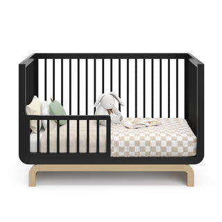 Baby crib in two-tone black and natural wood color in toddler bed conversion stage, with various decorative toddler bedroom items