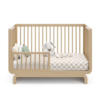Baby crib in natural wood color in toddler bed conversion stage, with various decorative toddler bedroom items