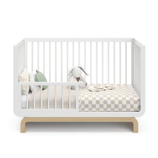 Baby crib in two-tone white and natural wood color in toddler bed conversion stage, with various decorative toddler bedroom items