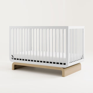 Baby crib in a two-tone white and natural wood color, situated at an angle in a studio setting
