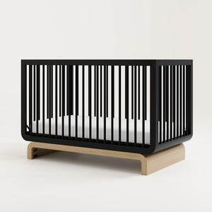 Baby crib in a two-tone black and natural wood color, situated at an angle in a studio setting
