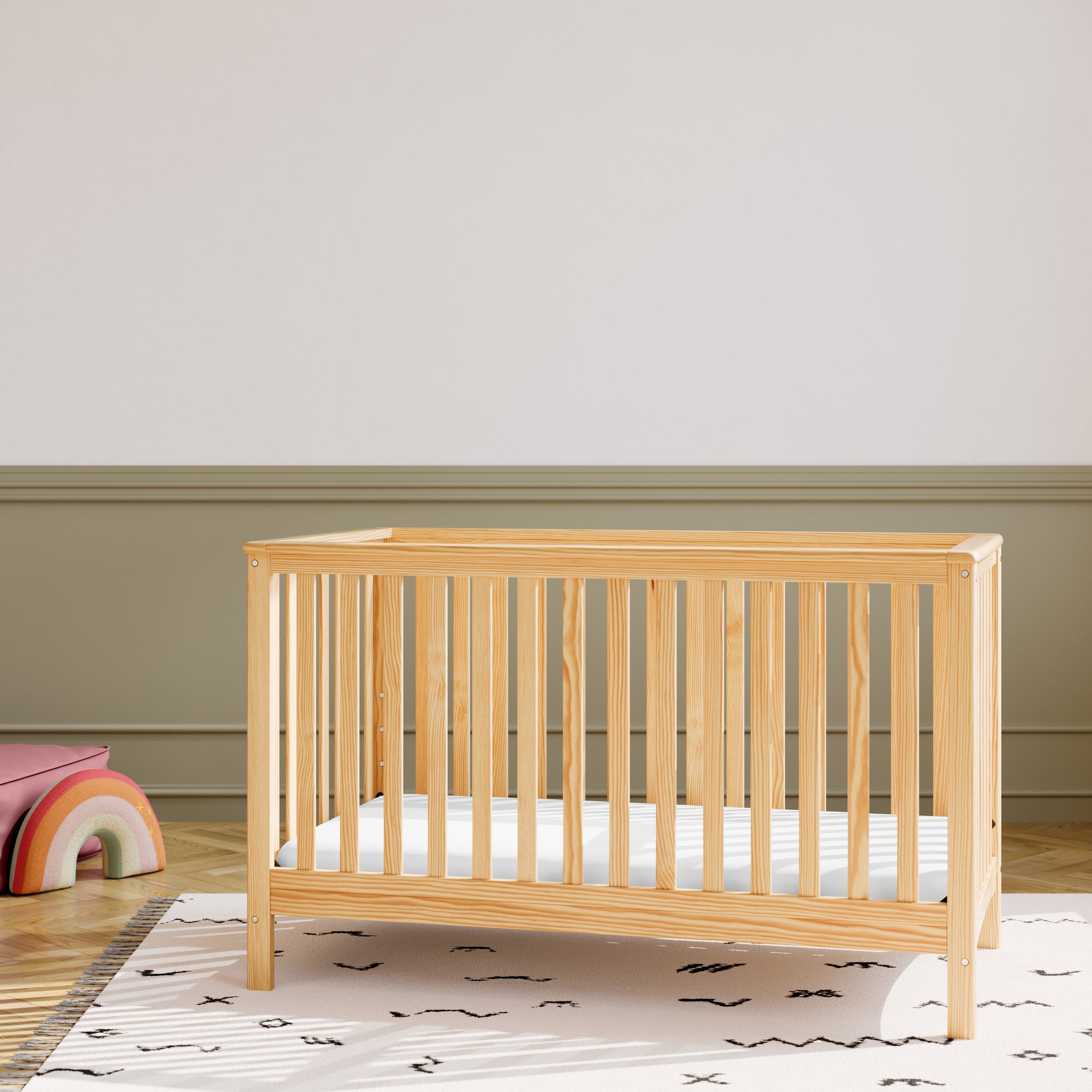 Natural wood baby crib in nursery room setting with olive green colored wall behind it and various decorative objects in the room, such as a rainbow plush toy
