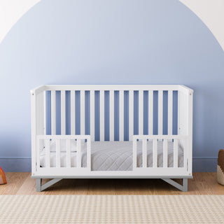 White crib with pebble gray in toddler bed conversion with two safety guardrails in nursery 